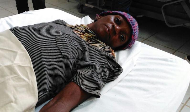 A Haitian woman lies on a hospital bed. She wears a striped shirt, a scarf, and a colorful headwrap.