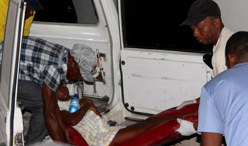 Three Haitian men carry a young woman out of an ambulance at night. Two hold her legs and one holds her torso. 