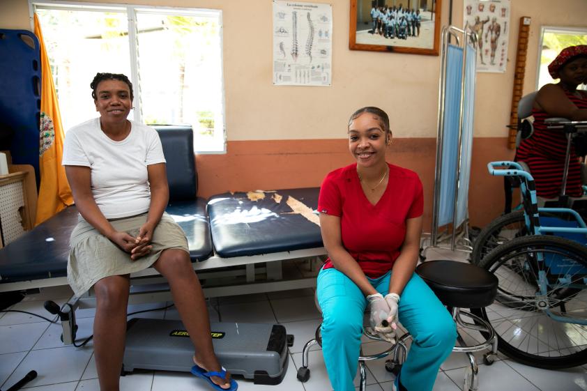 Two Haitian women sit in a brightly-lit rehab center, smiling warmly at the camera. One wears a white T-shirt and taupe skirt, and the other wears red and aqua scrubs and medical gloves.