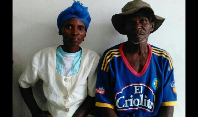 A Haitian couple sits side by side. The woman is on the left and wears a blue headwrap. The man is on the right and wears a large hat.