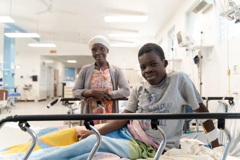A young Haitian boy props himself up in a hospital bed and smiles at the camera. His mother stands behind him, smiling.