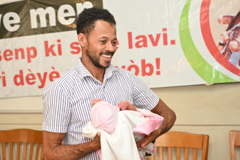 A light-skinned Haitian man in a short-sleeved button-down shirt stands in a hospital waiting room. He holds an infant in his arms and smiles joyfully.