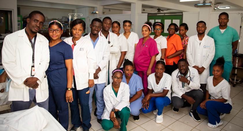 Group photo of 18 Haitian clinicians, men and women in scrubs or white lab coats, in a large white-and-green room.