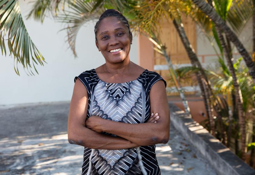 A Haitian woman in a black and white sleeveless dress stands under palm fronds. Her arms are crossed and she is smiling widely, her head slightly tilted.