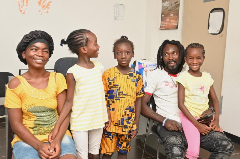 Three school-age children pose with their parents in a waiting room. The father and oldest child have caught each other's gaze and everyone is smiling happily.