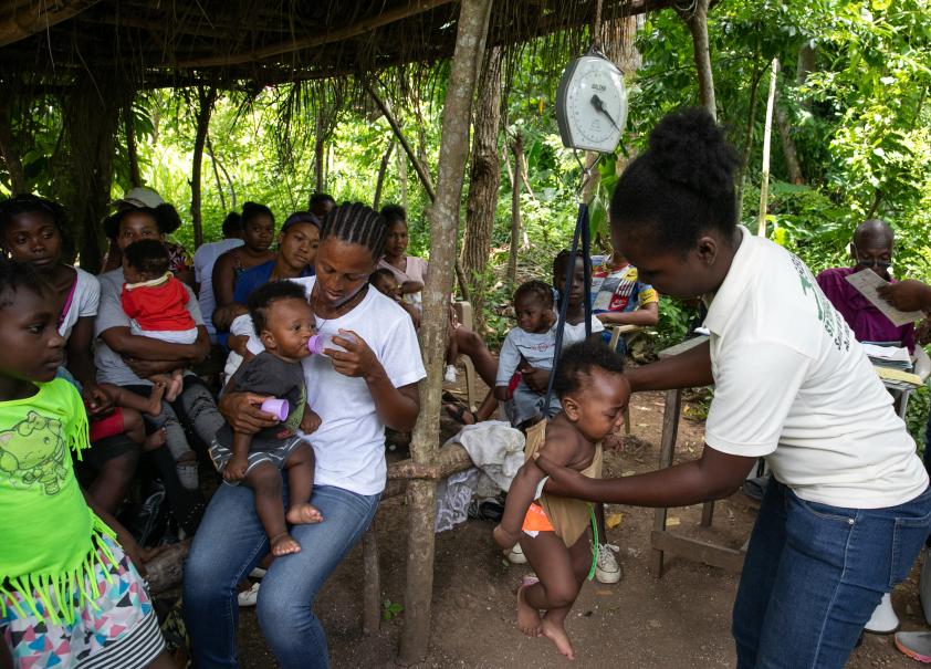 A Haitian woman in jeans and polo shirt weighs a baby using a hanging scale. Next to them, under a thatched roof, sit many mothers with their children.