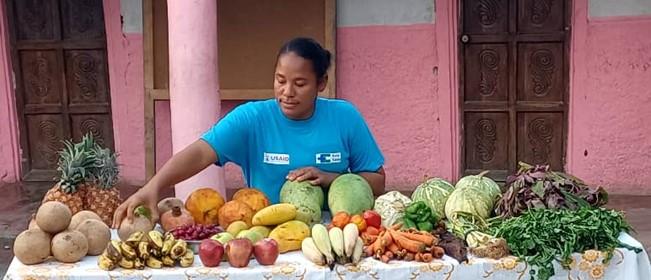 In front of a large pink building, a Haitian woman wearing a blue T-shirt with USAID and Kore Sante logos stands behind a large table loaded with 20 different kinds of fruits and vegetables.