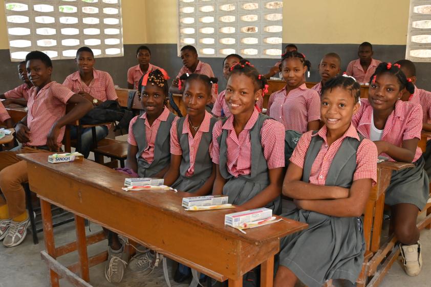 A dozen young Haitian teens in pink and gray school uniforms sit at desks with toothpaste and toothbrushes.