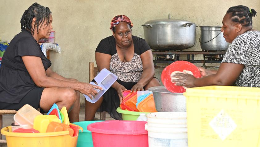 Three Haitian women sit on low chairs around large, colorful plastic basins, washing serving containers. Two large aluminum pots sit on a gas range behind them.