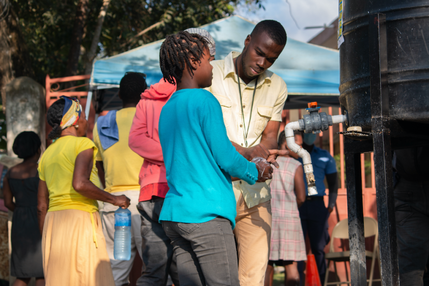 A Haitian man stands in front of a hand washing station in the community. He leans over and helps a young woman wash her hands with a small bar of soap.
