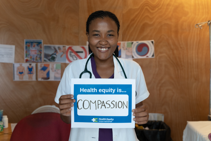 Dr. Catherine Scipion wears a white doctor's coat with a stethoscope draped around the back of her neck. She holds a white sign with a blue border that says "Health Equity is...Compassion"