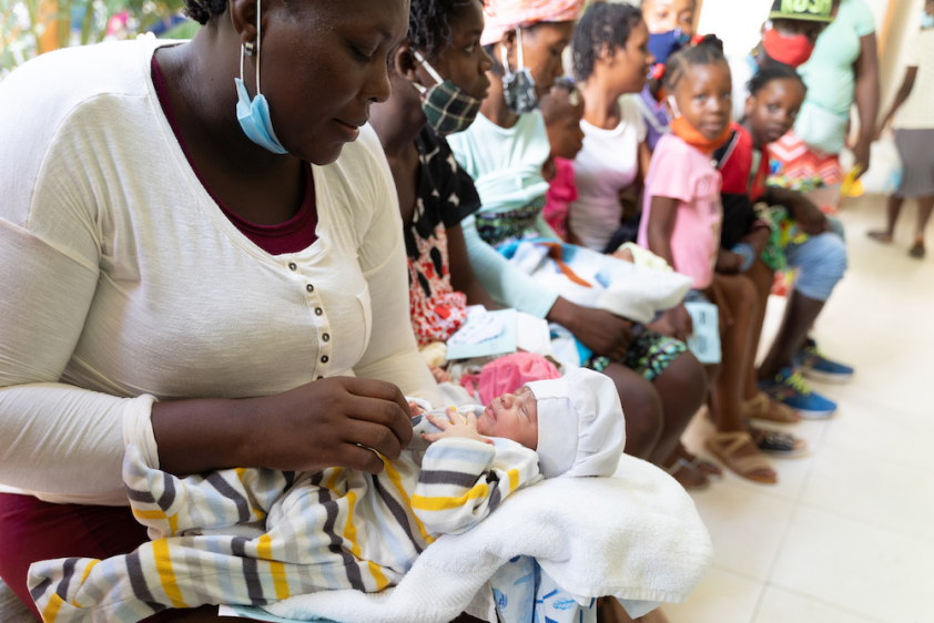 A mother sits with her baby in St. Boniface Hospital's courtyard. The mother wears a shite long sleeved shirt, with a blue medical mask under her chin. Her baby is in a yellow striped onesie with a white cap, laying on a blanket. There are many other patients sitting on the benches looking on.