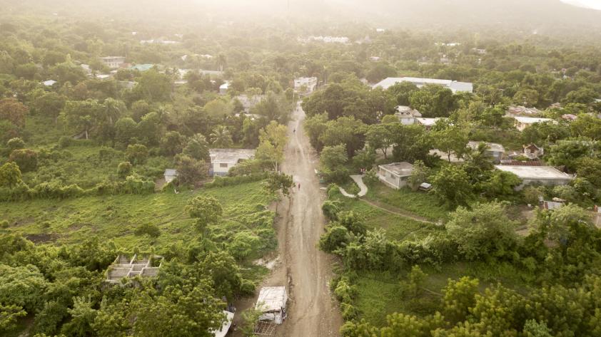 An arial view of a long dirt road surrounded on lush green fields on both sides.