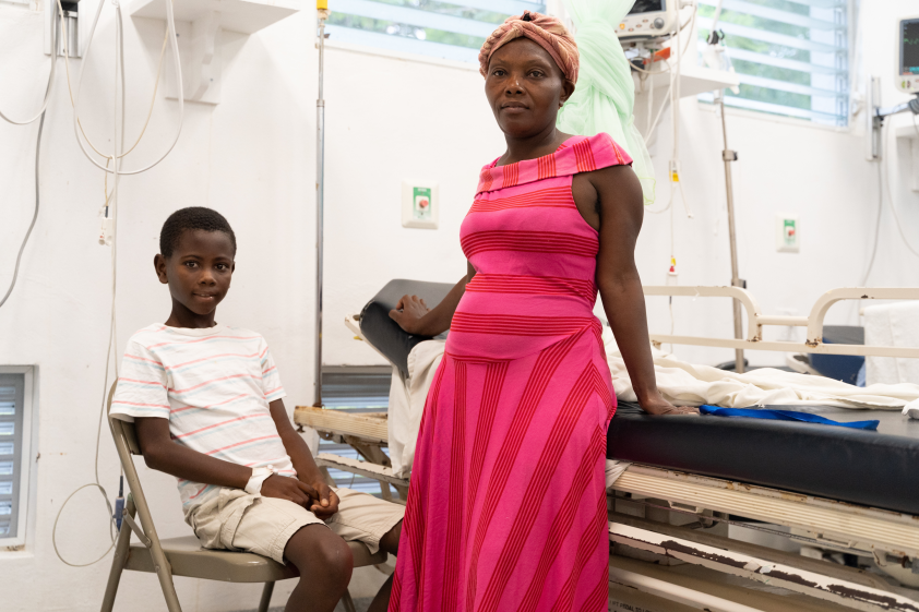 A young Haitian boy sits in a metal folding chair in a hospital ER. His mother stands to the right of him with her hand on an empty hospital bed.