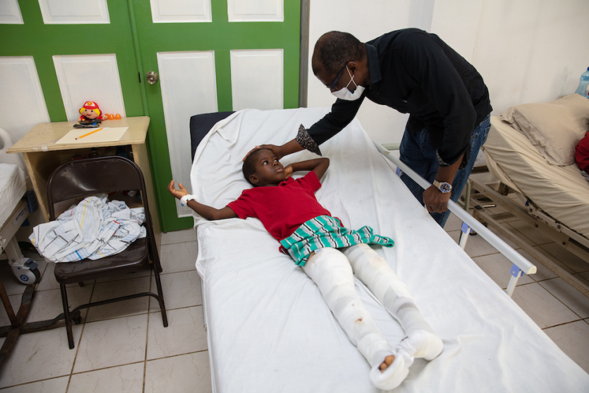 A yong boy lays in a hospital bed. He has two broken legs wrapped in white bandages. A doctor leans over and touches the boy's head.