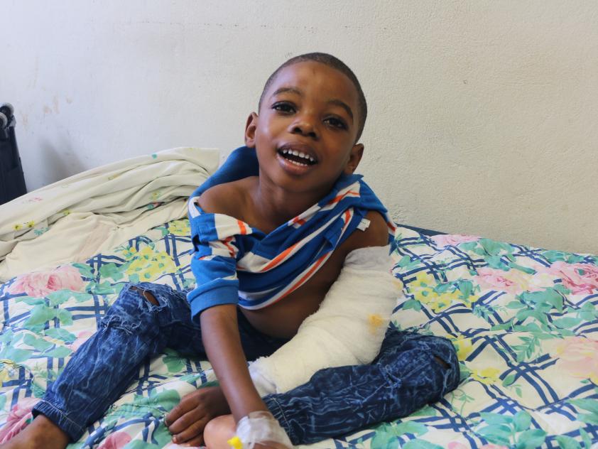 A young Haitian boy sits on a hospital bed. His arm is covered in a cast up to his elbow.