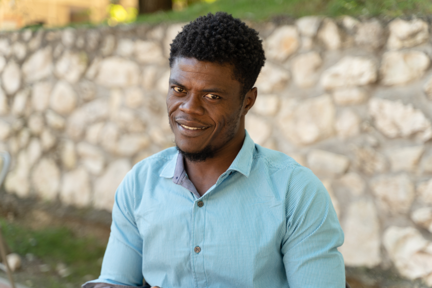 A Haitian man sits in front of a low stone wall. He wears a light blue button down shirt and has short, dark hair. He smiles directly at the camera.