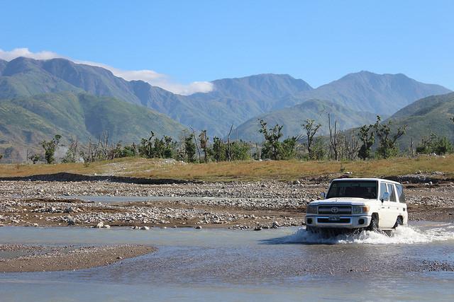 A white ambulance drives through a shallow river. There is a grassy plain and mountains in the background.