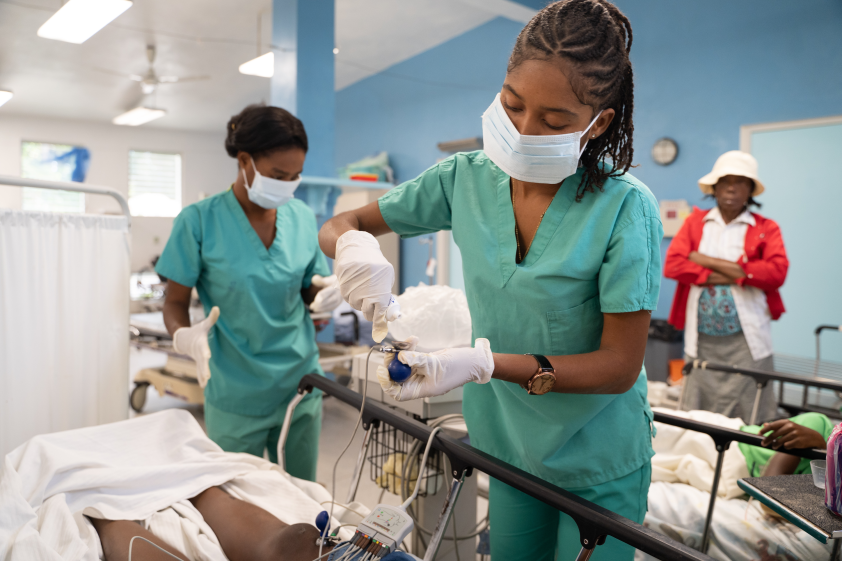 A female Haitian clinician wearing light blue scrubs and a face mask leans over a hospital bed to adjust medical equipment for a patient.