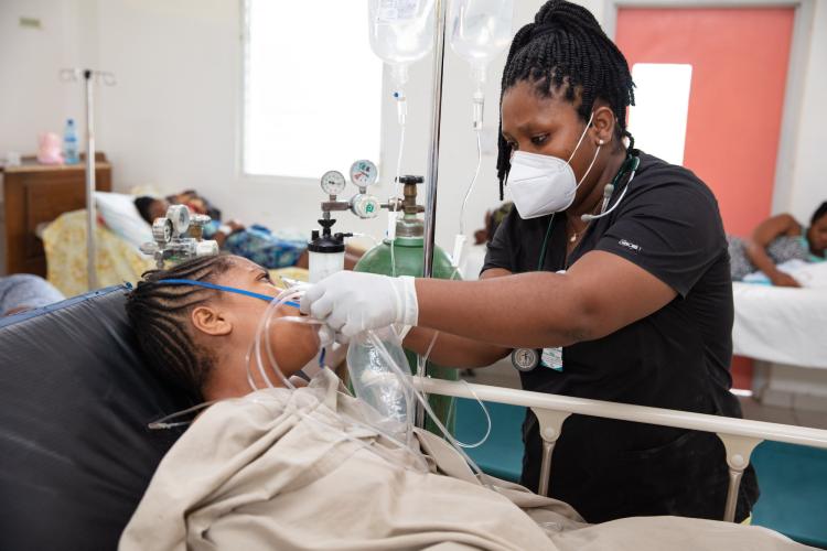 A woman patient lays in a hospital bed with a blanket covering her torso as a woman Haitian doctor puts an oxygen mask to her face. The doctor stands to the right of the bed. The doctor wears a white medical mask, black scrub top, and white exam gloves. Her hair is intricately braided. An IV pole stands to the left of the doctor, and to the left of the IV is a large green oxygen cylinder.