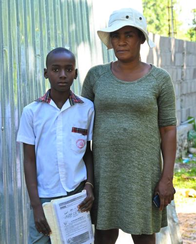 A Haitian woman in gray dress & white hat stands protectively next to her 10-year-old son, who wears a crisp white shirt.gray dress and white hat stands protectively next to her 10-year-old son, who wears a crisp white shirt.