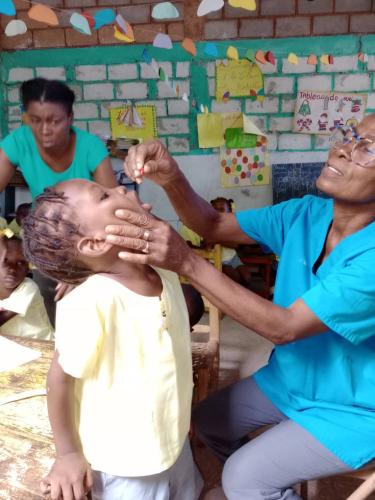 Inside a cinderblock classroom decorated with brightly-colored paper, a Haitian woman in glasses and blue scrubs squeezes a capsule into a preschool-age girl’s mouth.