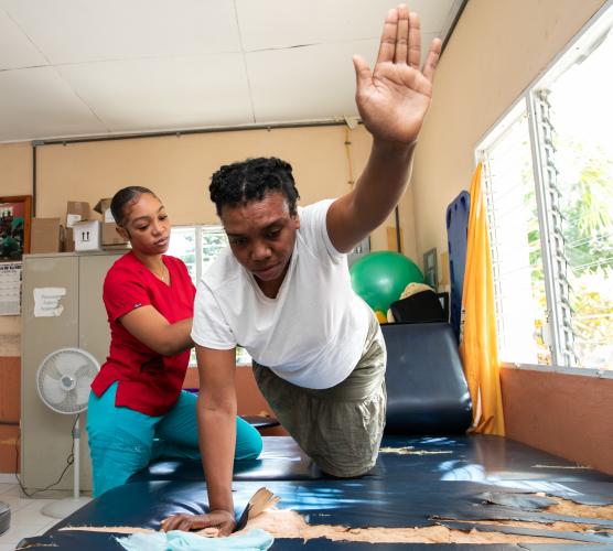 A woman in a white T-shirt balances on a well-worn rehab table. She stretches her left hand out in front of her while a therapist helps support and stretch her right leg.