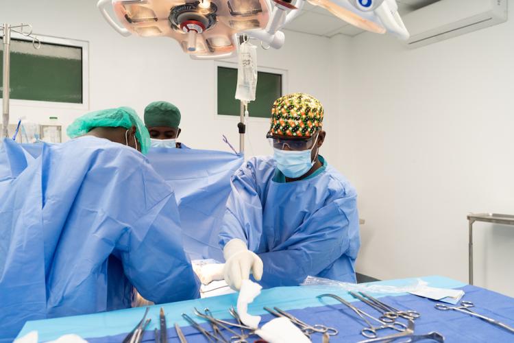 Three surgeons perform a procedure. They all wear light blue surgical gowns, caps, and surgical masks. A table of equipment stands in the foreground.