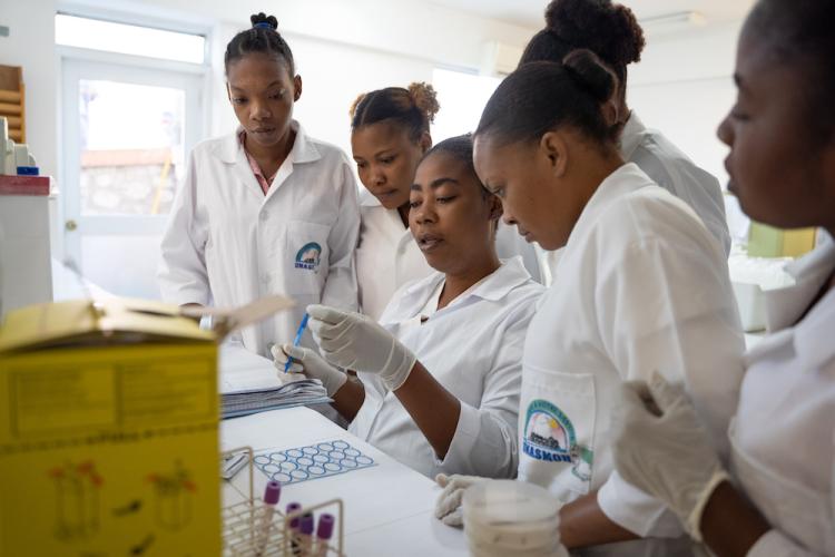 A group of women Haitian medical students stand around a seated woman lab technician and observe her work.