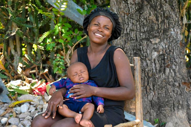 A Haitian woman sits outside in a wooden chair near a large tree trunk. She holds her infant son in her lap. He wears blue shorts and a plaid button down-shirt. He is sleeping. The woman wears a black hair bonnet and a sleeveless black dress. She has a bright smile.