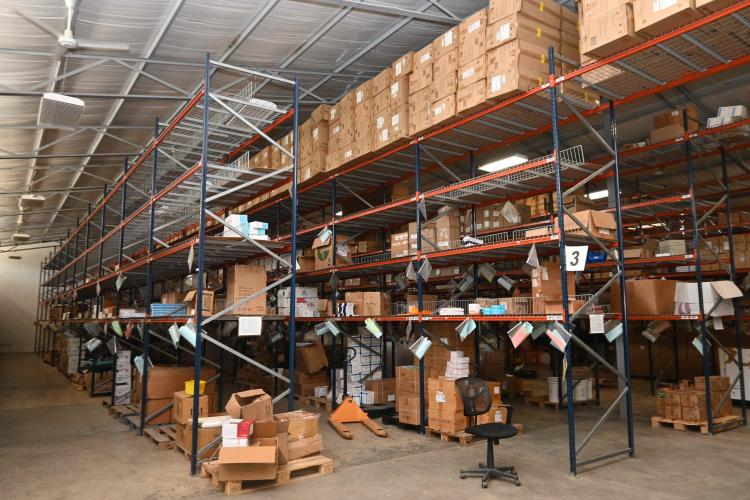 The inside of a cavernous warehouse with cardboard boxes on pallets and long rows of shelving, and colored tags on the shelves.