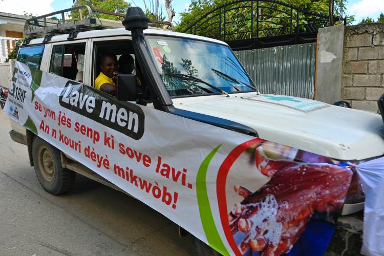 A white 4x4 vehicle, handwashing banner in Haitian Creole hanging on the side, drives down a village street.