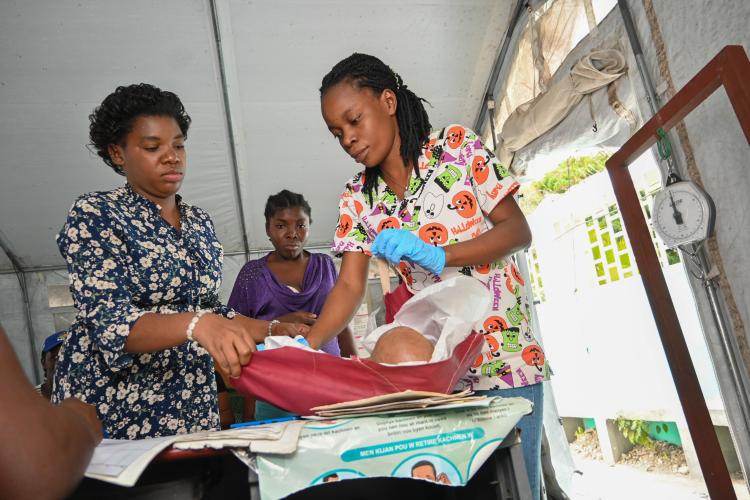 A Haitian nurse lays a baby on a red cloth to prepare the child for weighing. A woman with short hair and a printed button-down shirt stands to the left assisting the nurse.