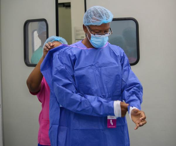 Assisted by a woman in pink scrubs, a Haitian man wearing glasses, surgical mask, and head covering adjusts the sleeves of a blue surgical gown.