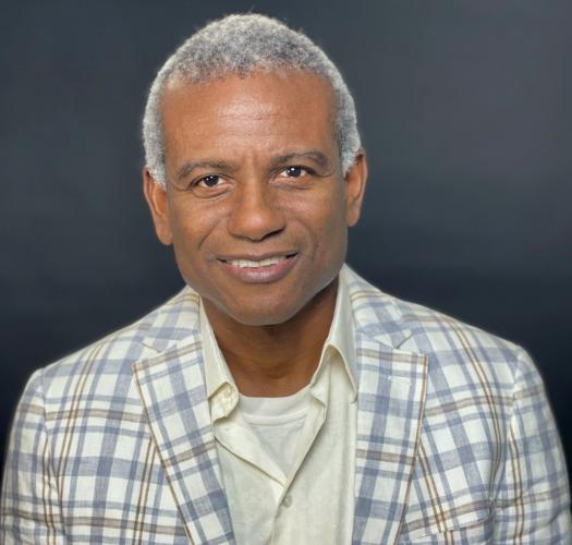 Headshot of a smiling Haitian man with short-cropped gray hair, wearing a light-colored plaid jacket over a white shirt.