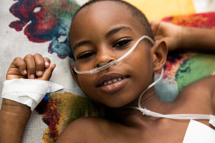 Close-up portrait of a young Haitian boy half-smiling at the camera. As he relaxes on a patterned pillow, his hands by his head, tubes bring oxygen to his nostrils.