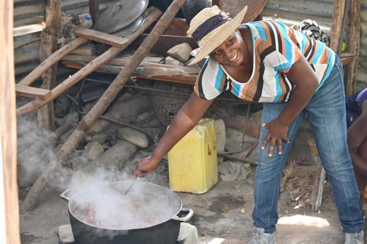 A woman wearing a bright striped shirt and a stray hat smiles as she leans over to stir a large pot.