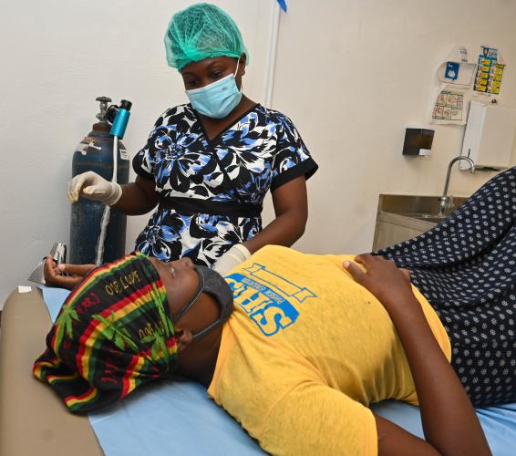 A Haitian woman in gold T-shirt lies on an exam table. She makes eye contact with a nurse who smiles behind her face mask.
