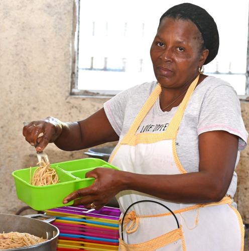 A middle-aged Haitian woman looks over at the camera as she fills a serving container with spaghetti. She is wearing a black hair net, pale T-shirt, and white apron.