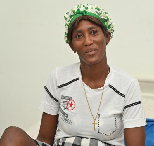 Portrait of a Haitian woman smiling gratefully at the camera. She wears a green head wrap, a white knit shirt, and a large gold cross.