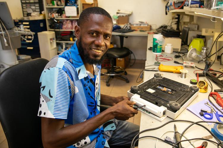 A Haitian biomedical techniican smiles as he works on repairing medical equipment. He is in a repair shop filled with wires, equipment, and tools.