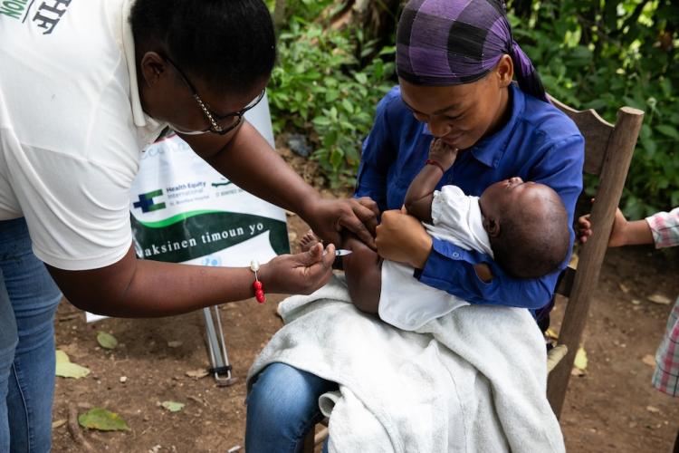 A Haitian mother holds her baby as a Haitian nurse vaccinates the baby in the thigh.