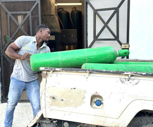 A Haitian man in gray polo shirt carefully slides a green oxygen cylinder into the bed of a pickup truck. Large black tanks with hoses can be seen in the room behind him.