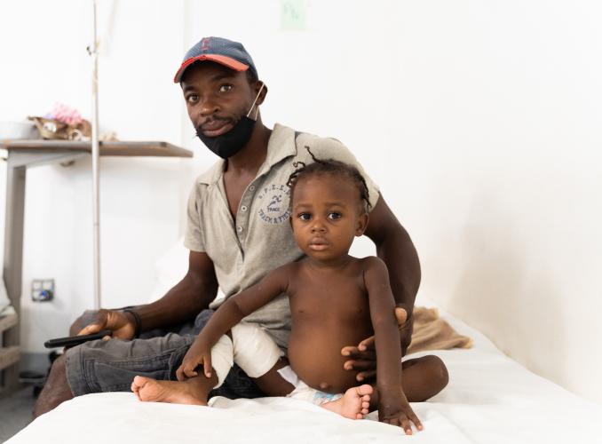 A small toddler with bandaged leg and diaper sits on a white hospital bed. Her father wraps a protective arm around her.