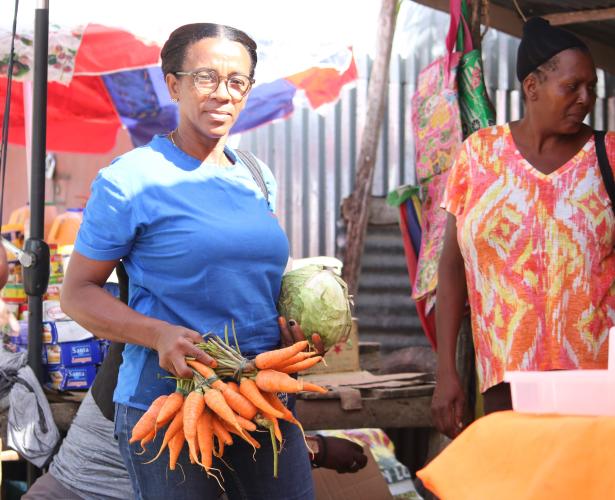 Two Haitian women stand in a busy outdoor market, in front of colorful product displays. One holds several bunches of carrots and a cabbage.