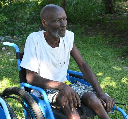 ¾ portrait of an older Haitian man sitting in a blue wheelchair, looking into the distance. He is lean and wiry, with a close-cropped white beard.