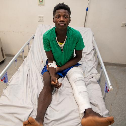 A young Haitian man sits on a hospital gurney, his left leg fully bandaged in white. He is wearing blue shorts and a green short-sleeved shirt.