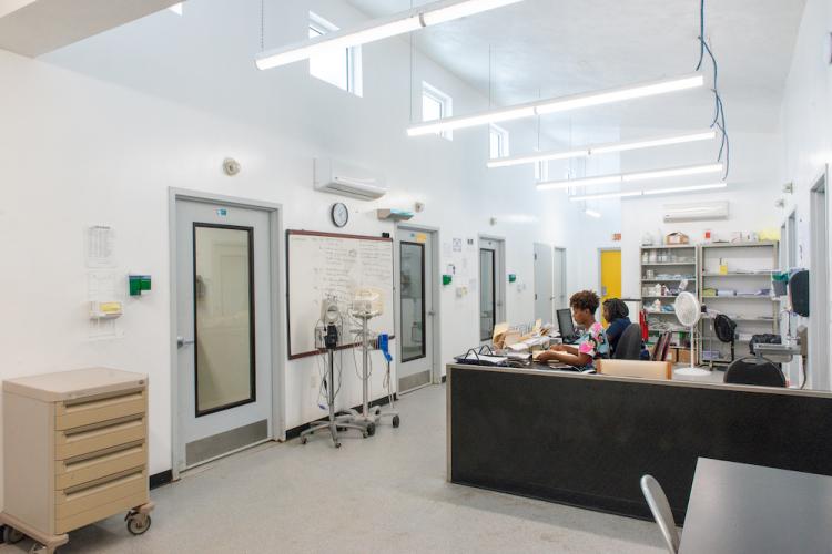 A panoramic view of an infectious disease ward: two nurses sit a long L-shaped desk and face a wall with three doors. A whiteboard hangs on the wall.