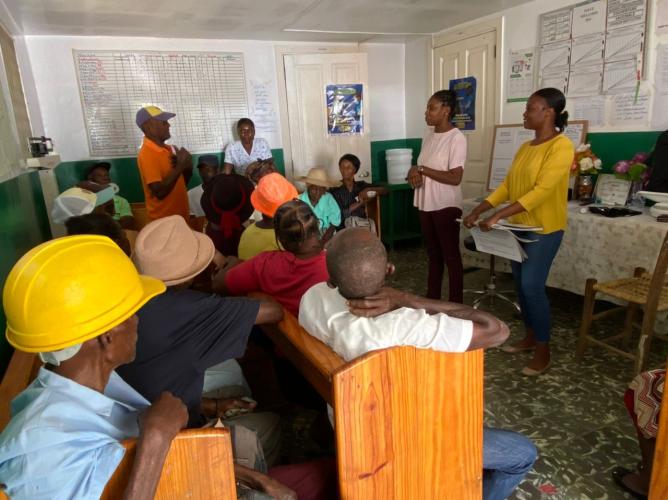 Two women community health workers lead a training in rural Haiti. The two women stand in front of the room. Their students sit on long benches in front of them in a small room.