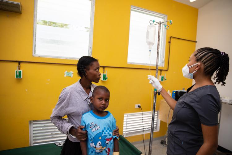 A young Haitian boy stands in front of his mother in our infectious disease center. A nurse stands to the right and adjusts and IV pole.